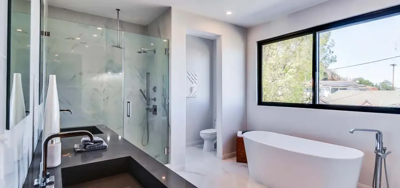 Modern minimalist bathroom with freestanding white bathtub near large window overlooking trees, glass-enclosed shower with marble-like tile, and sleek dark countertop with chrome fixtures.