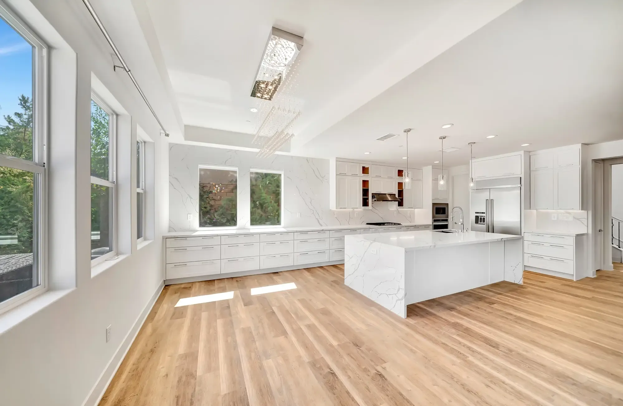 A spacious modern kitchen with white marble island, white cabinetry, stainless steel appliances, and natural light from windows