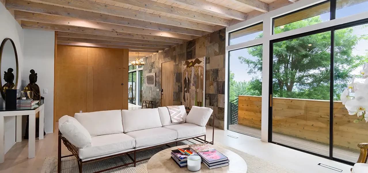 A modern living room with white sofa, exposed wooden beams, floor-to-ceiling windows, and decorative Buddha statues